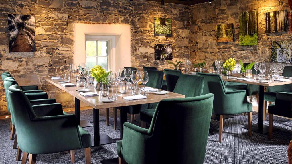 Elegant dining room with green velvet chairs, stone walls, soft lighting, and nature artwork, perfect for a cosy meal.