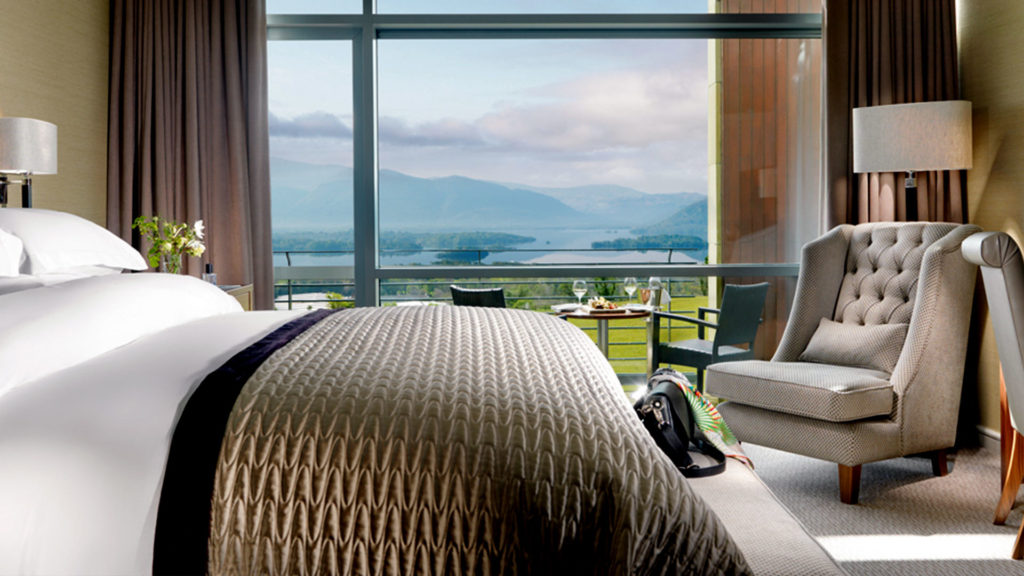 Luxurious hotel room with plush bedding, armchair, and large window offering breathtaking lake and mountain views.