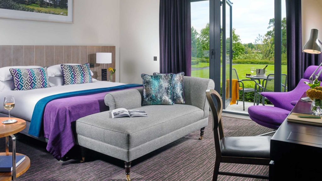 Modern hotel room with plush bed, chaise longue, patio doors opening to lush garden, cosy seating, and sophisticated decor.