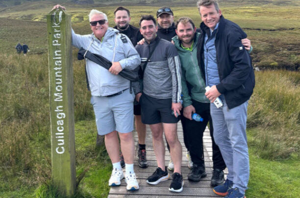Six men pose and smile together on a wooden boardwalk next to a sign reading 