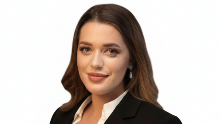 A woman with long brown hair, wearing a black blazer and white blouse, poses against a plain white background.